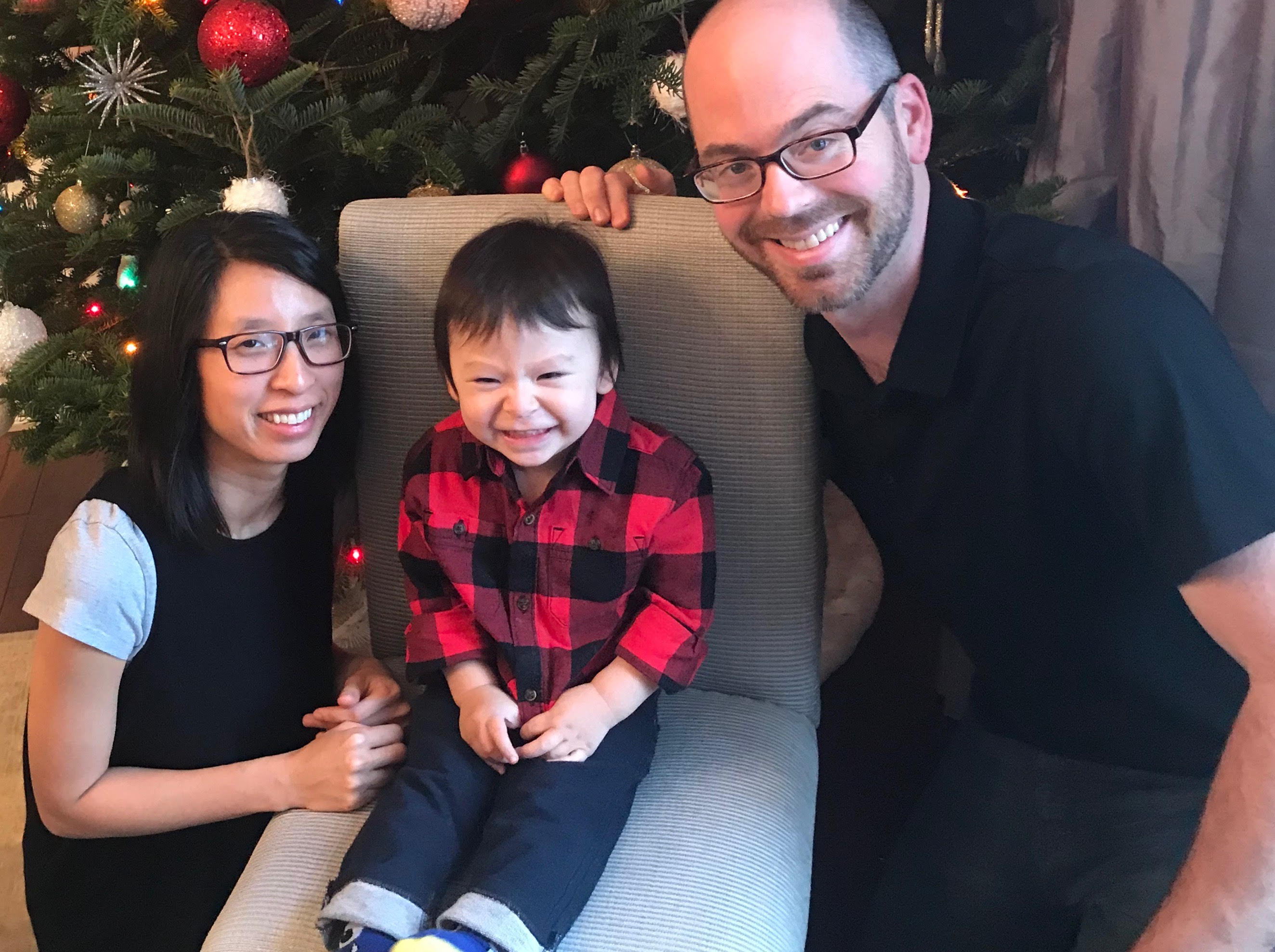 A young family smiles in front of a Christmas tree